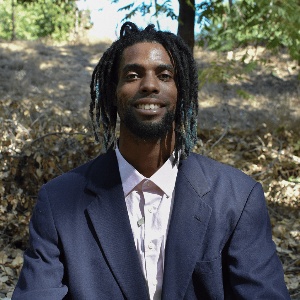KamRon Perry, SF Bay Area creative professional, smiles warmly while seated outdoors. Wearing a dark suit jacket over a light pink shirt, his dreadlocks and genuine expression convey approachable confidence.
