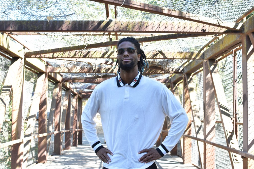 KamRon Perry, SF Bay Area creative professional, poses confidently on a metal bridge wearing a white polo and sunglasses. Clean urban aesthetic meets natural light.